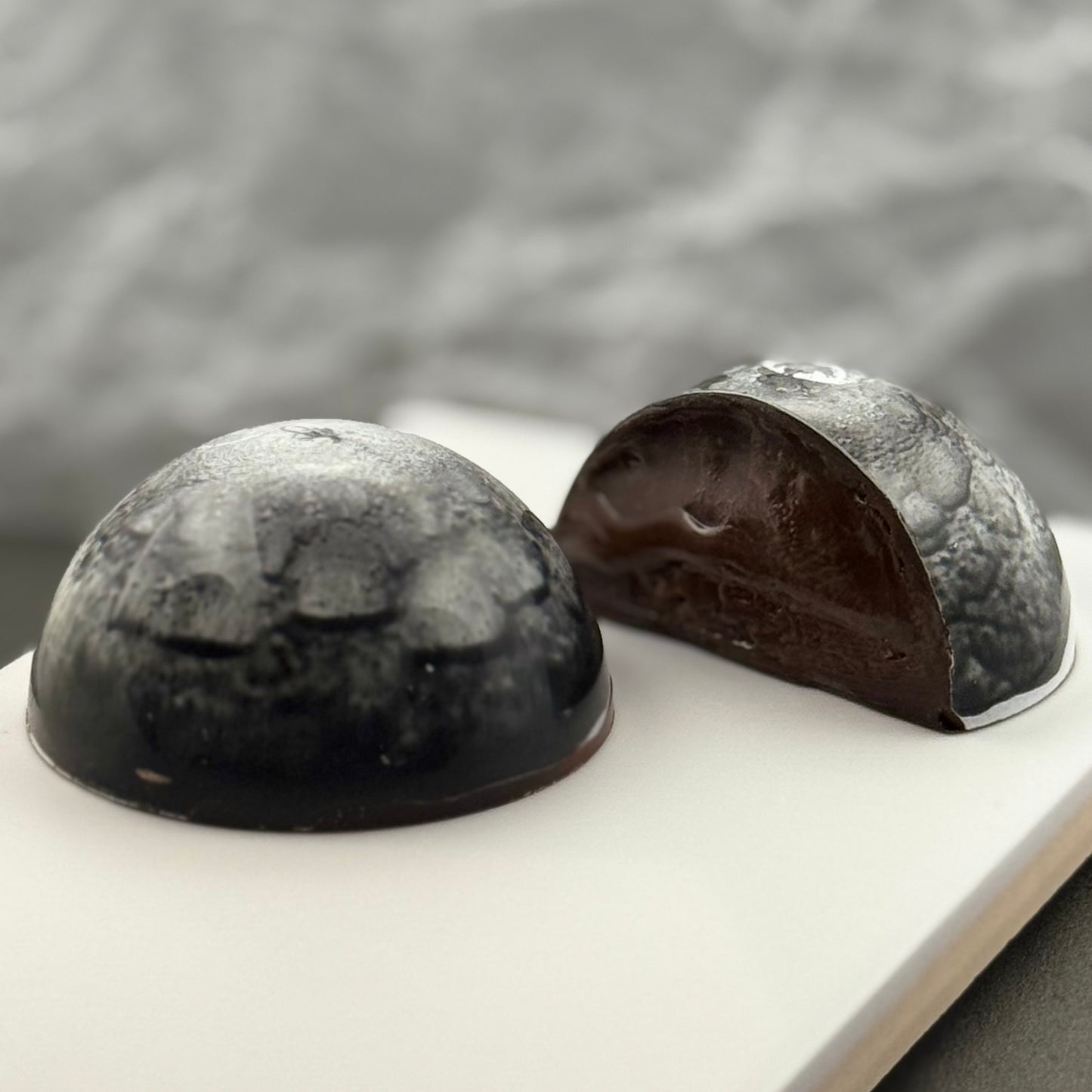 A photo of two bonbons, one whole and one cut in half to show the salted caramel ganache and the almond praline layer, with a moody dark shell.